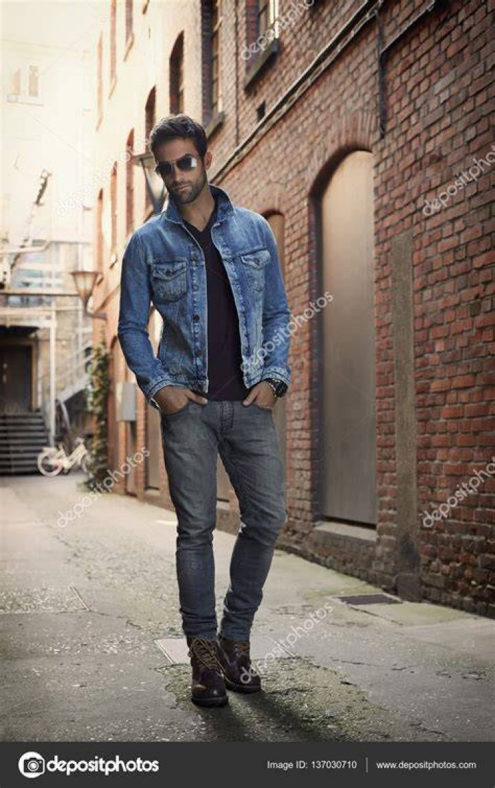 a man wearing a denim jacket and studded shirt looks at the camera with